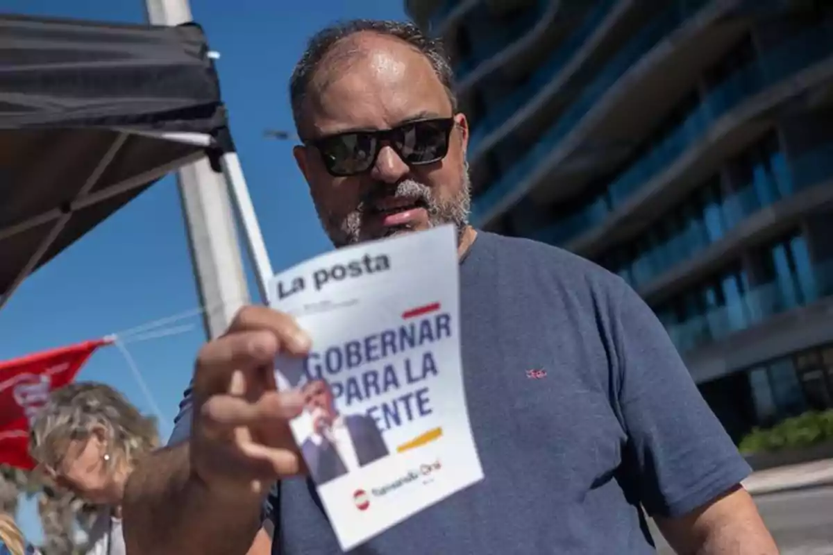 Hombre con gafas de sol sosteniendo un folleto de campaña política en la calle frente a un edificio moderno Hombre con gafas de sol sosteniendo un folleto de campaña política en la calle frente a un edificio moderno