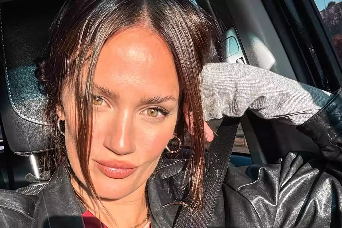 Mujer joven con cabello castaño y ojos claros sentada en un automóvil usando chaqueta de cuero y suéter gris mientras mira a la cámara