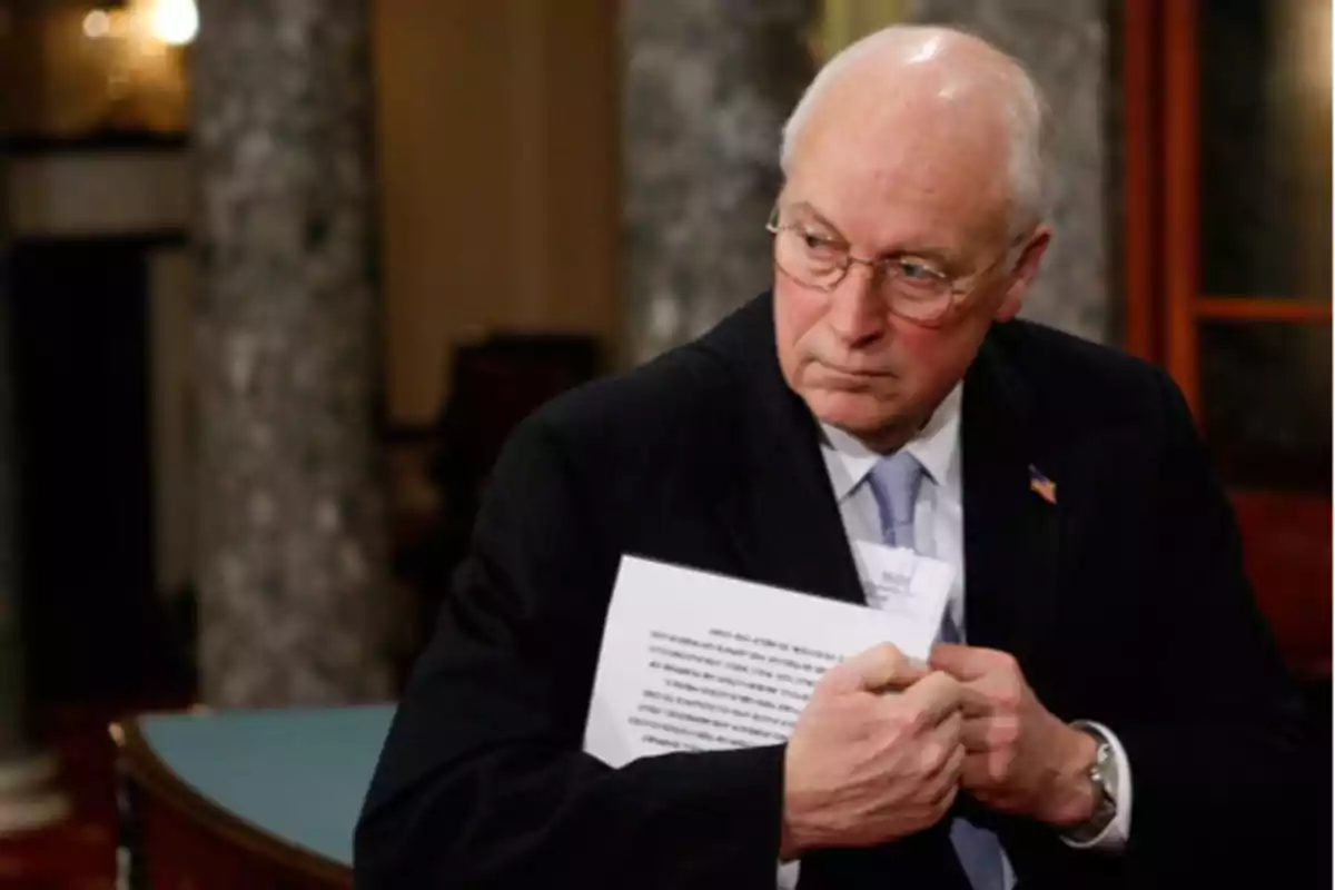 An older man with glasses and a dark suit holds some documents in a formal setting. An older man with glasses and a dark suit holds some documents in a formal setting.