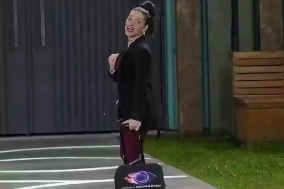 A person with a high bun and dressed in black walks carrying a suitcase with the Big Brother logo in an outdoor setting. A person with a high bun and dressed in black walks carrying a suitcase with the Big Brother logo in an outdoor setting.