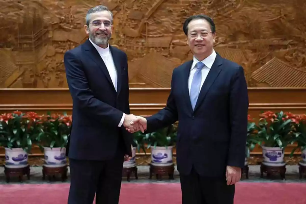 Two men in suits shake hands in a formal setting with a decorative background and plants.