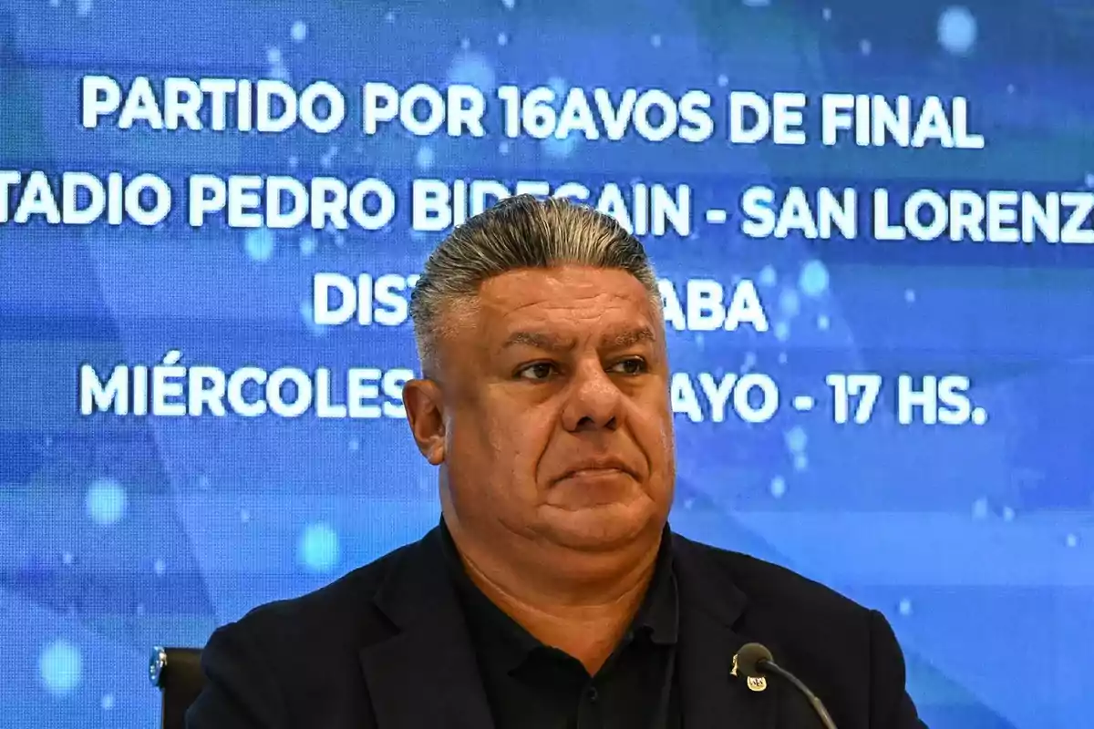 Hombre de cabello canoso y expresión seria frente a una pantalla azul con texto sobre un partido de fútbol en el estadio Pedro Bidegain Hombre de cabello canoso y expresión seria frente a una pantalla azul con texto sobre un partido de fútbol en el estadio Pedro Bidegain