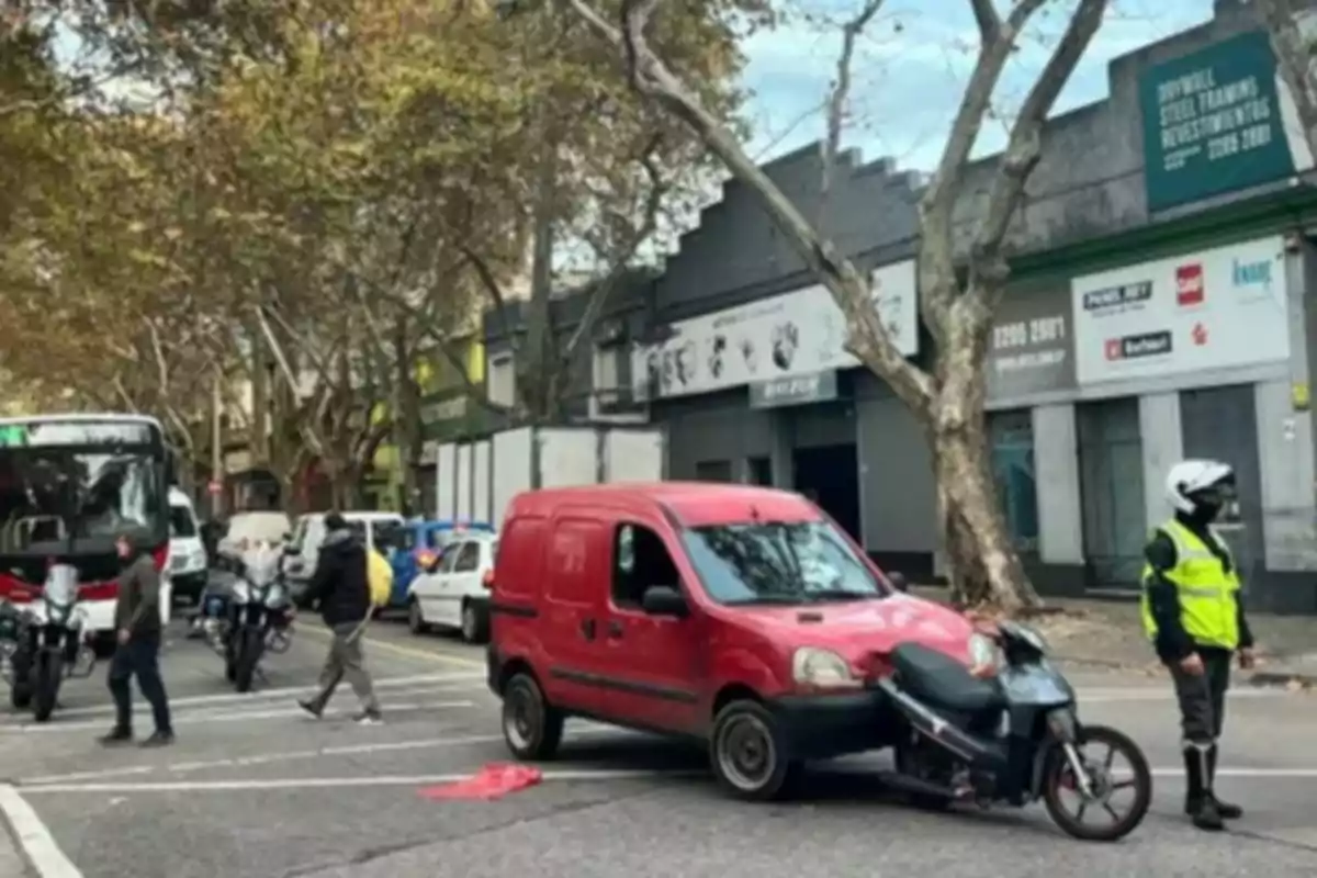 A red pickup truck crashed into a black motorcycle on an urban street while a traffic police officer observes the scene and several people and vehicles are around.