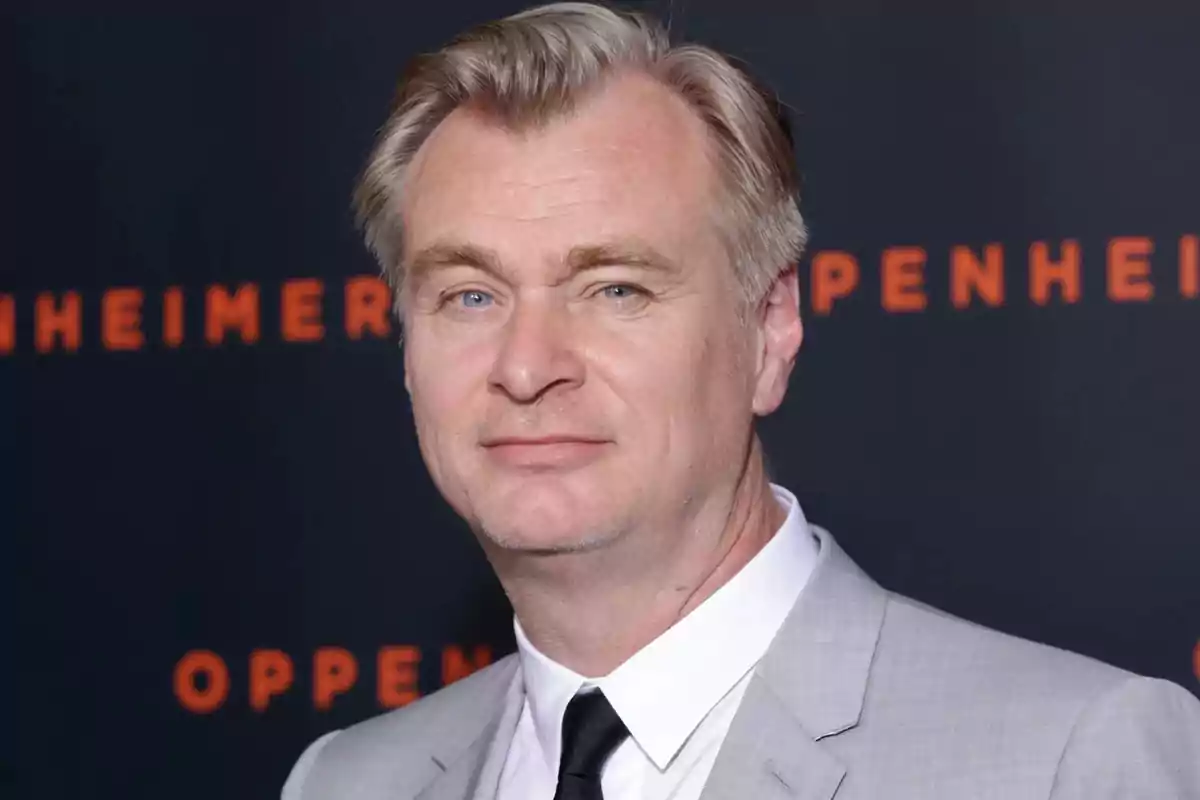 A man in a gray suit and black tie poses in front of a dark background with orange letters. A man in a gray suit and black tie poses in front of a dark background with orange letters.