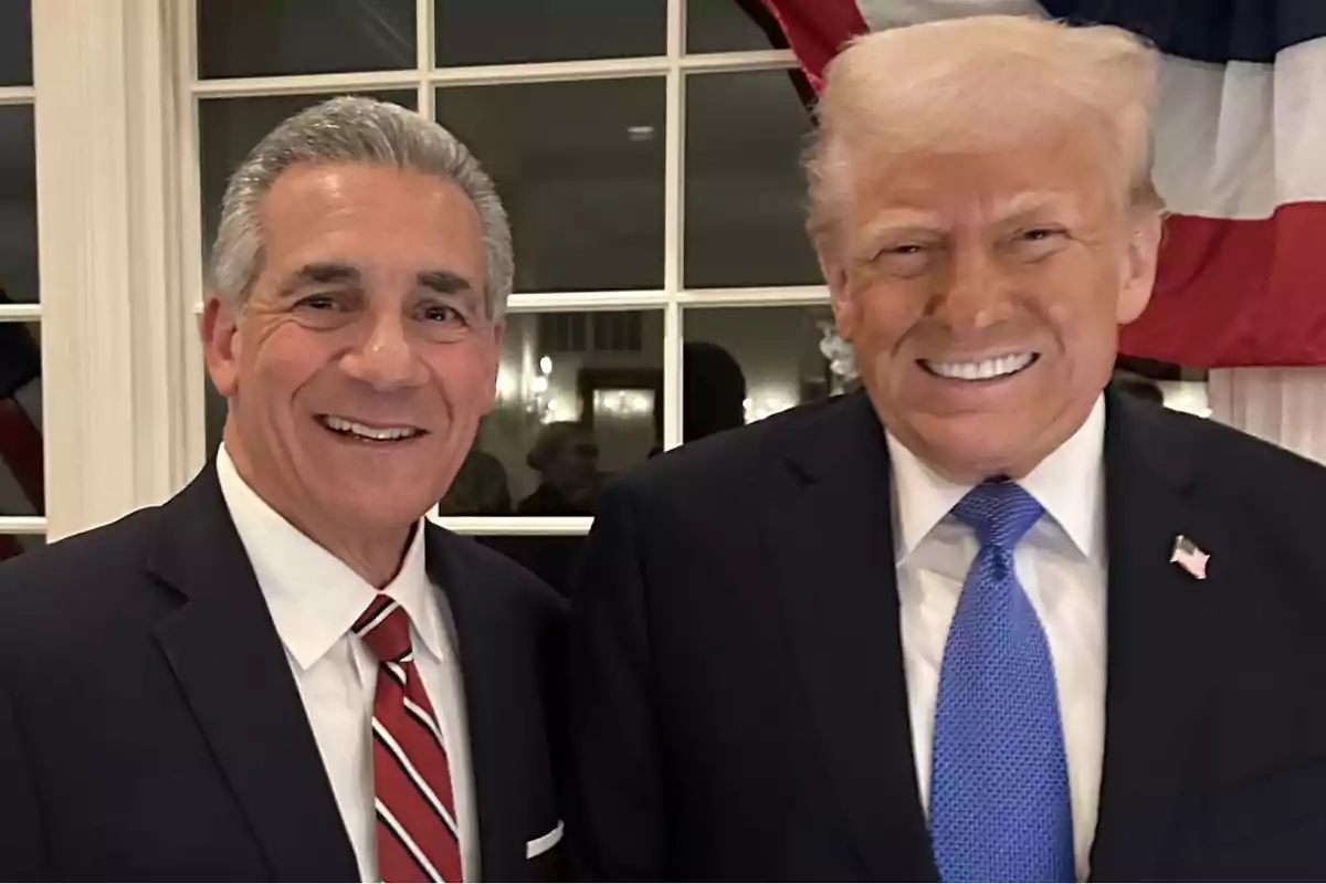 Dos hombres sonrientes posan juntos en un evento formal con una bandera de fondo.