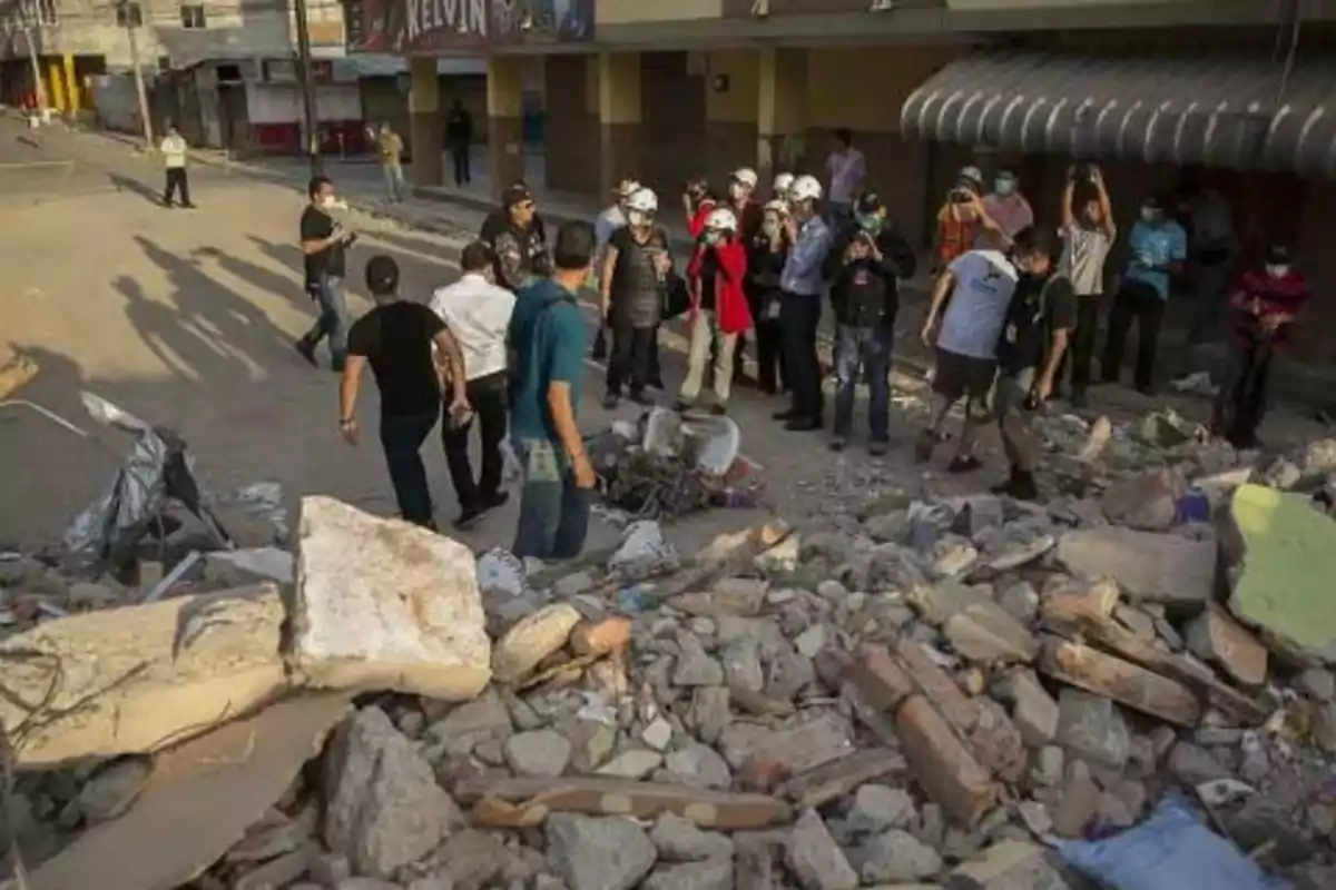 People look at the rubble of a collapsed building on a street after a natural disaster.