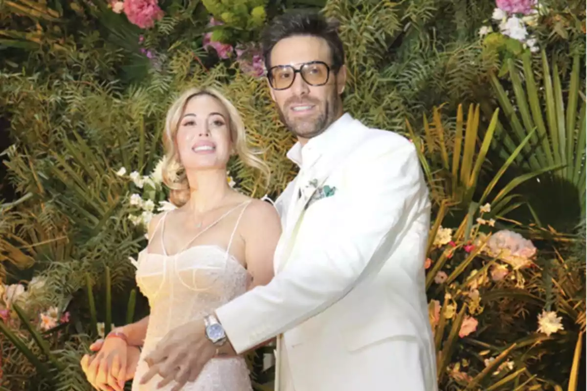 Una pareja vestida de blanco posa frente a un fondo de vegetación y flores.