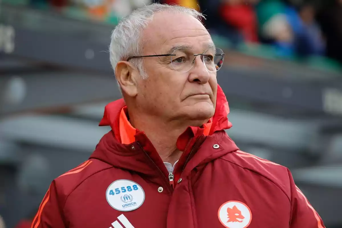 Hombre mayor con gafas y cabello canoso vistiendo una chaqueta deportiva roja con insignias y mirando hacia un lado en un entorno al aire libre Hombre mayor con gafas y cabello canoso vistiendo una chaqueta deportiva roja con insignias y mirando hacia un lado en un entorno al aire libre