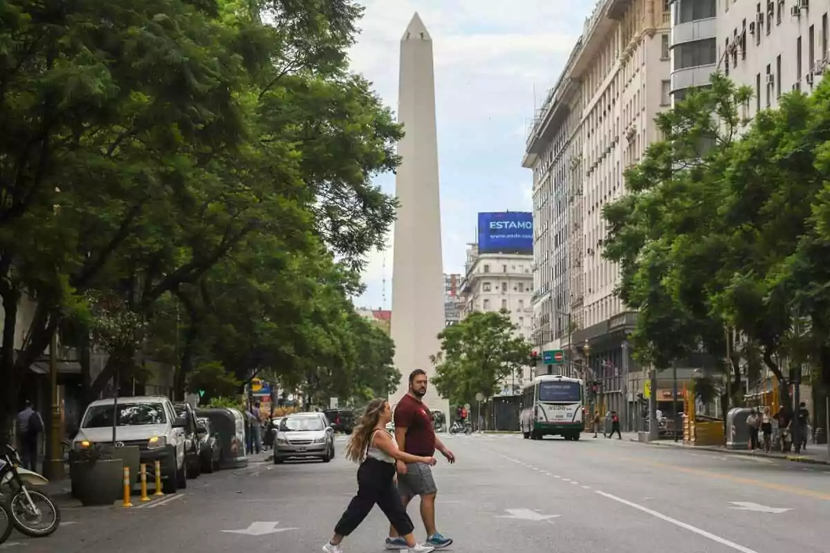 Una pareja cruza una calle en una ciudad con un obelisco al fondo rodeado de edificios y árboles. Una pareja cruza una calle en una ciudad con un obelisco al fondo rodeado de edificios y árboles.