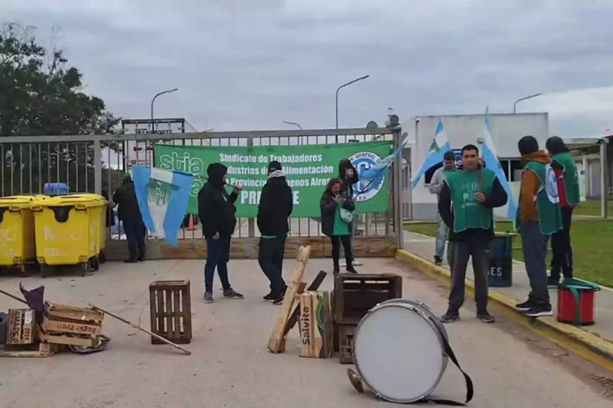 Un grupo de personas con chalecos verdes y banderas se encuentra frente a una reja con un cartel del sindicato de trabajadores de la alimentación, junto a contenedores amarillos y algunos objetos en el suelo.