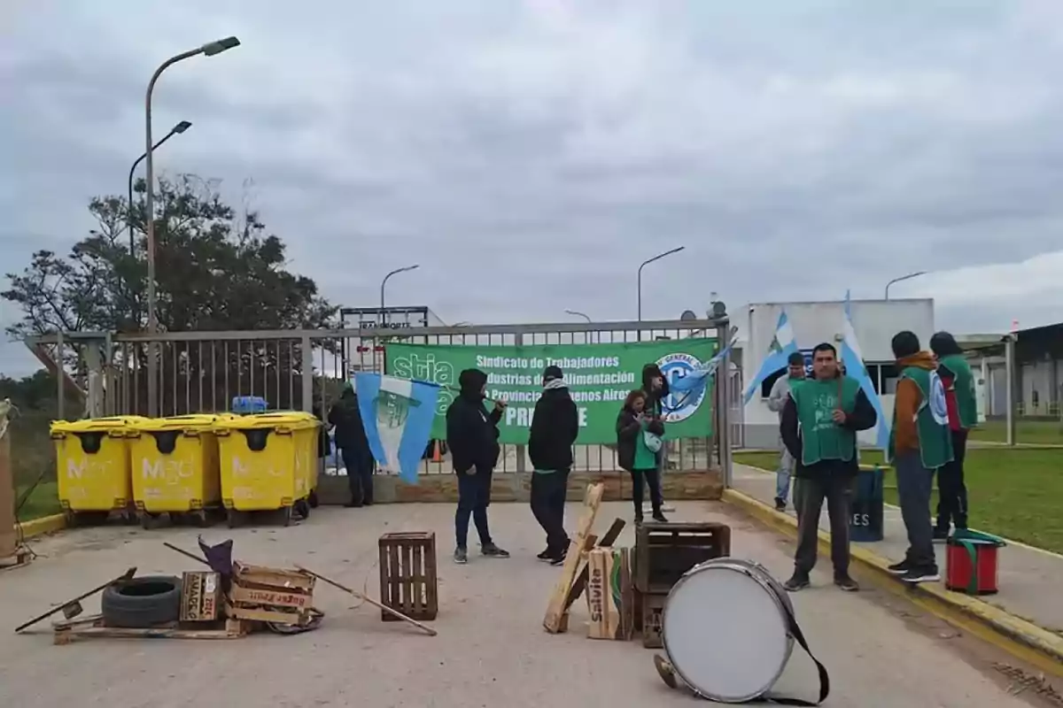 Personas con chalecos verdes y banderas argentinas se encuentran frente a una reja con un cartel verde que dice "Sindicato de Trabajadores de Industrias de la Alimentación de la Provincia de Buenos Aires" y varios contenedores amarillos.