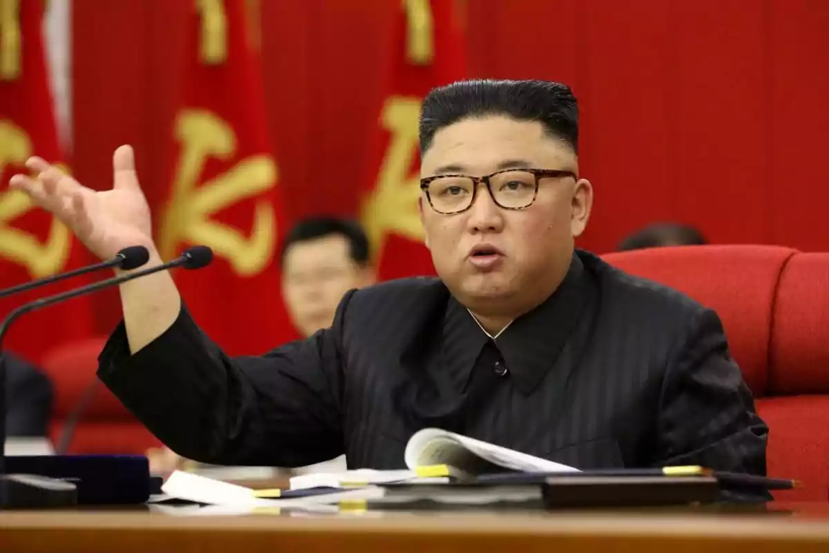 Hombre con gafas y traje oscuro hablando en una reunión con banderas rojas al fondo Hombre con gafas y traje oscuro hablando en una reunión con banderas rojas al fondo