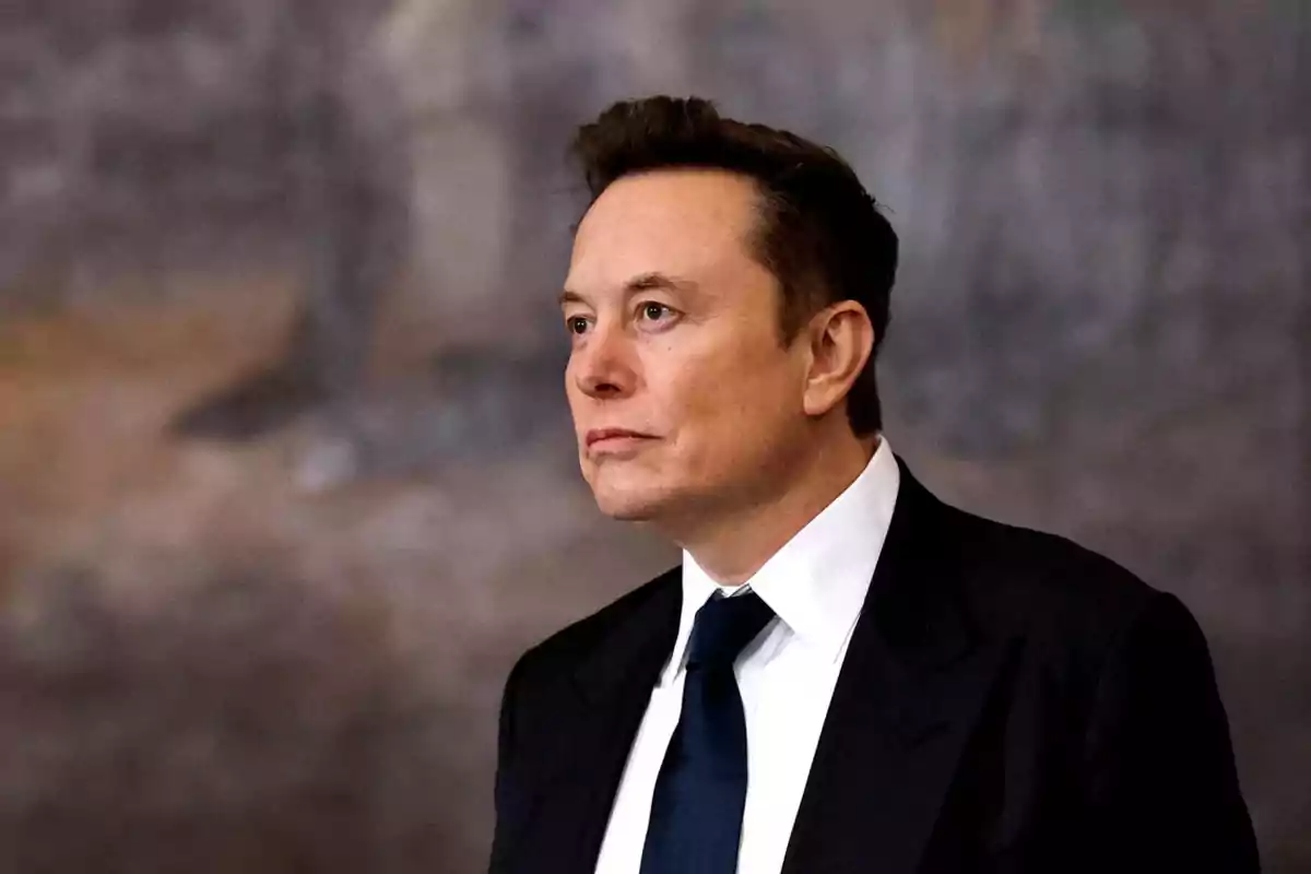 Short dark-haired man wearing a black suit with a white jersey and blue tie looking to the right with a serious expression against a blurred background.
