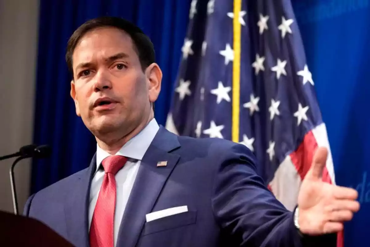 A man in a blue suit with a red tie speaking in front of a microphone with a flag in the background. A man in a blue suit with a red tie speaking in front of a microphone with a flag in the background.