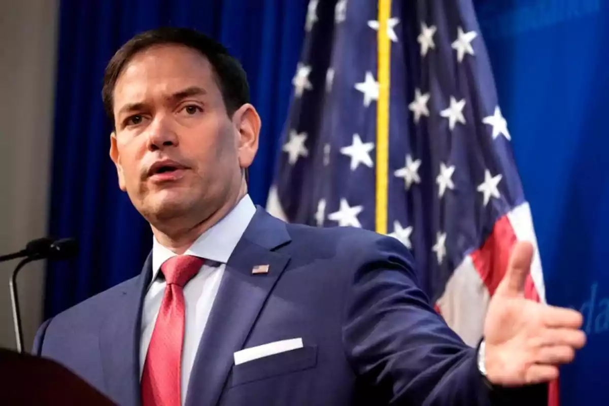A man in a blue suit with a red tie gestures while speaking in front of a microphone with a United States flag in the background. A man in a blue suit with a red tie gestures while speaking in front of a microphone with a United States flag in the background.