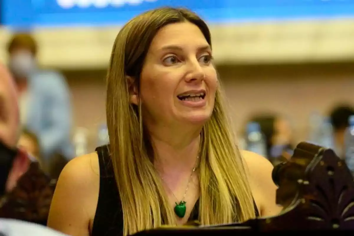 A woman with long blonde hair is speaking in what appears to be a meeting or conference setting, wearing a necklace with a green pendant. A woman with long blonde hair is speaking in what appears to be a meeting or conference setting, wearing a necklace with a green pendant.