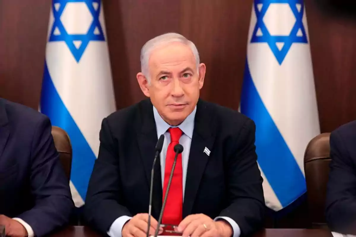 Un hombre de cabello canoso y traje oscuro con corbata roja está sentado frente a dos micrófonos, con banderas de Israel en el fondo. Un hombre de cabello canoso y traje oscuro con corbata roja está sentado frente a dos micrófonos, con banderas de Israel en el fondo.