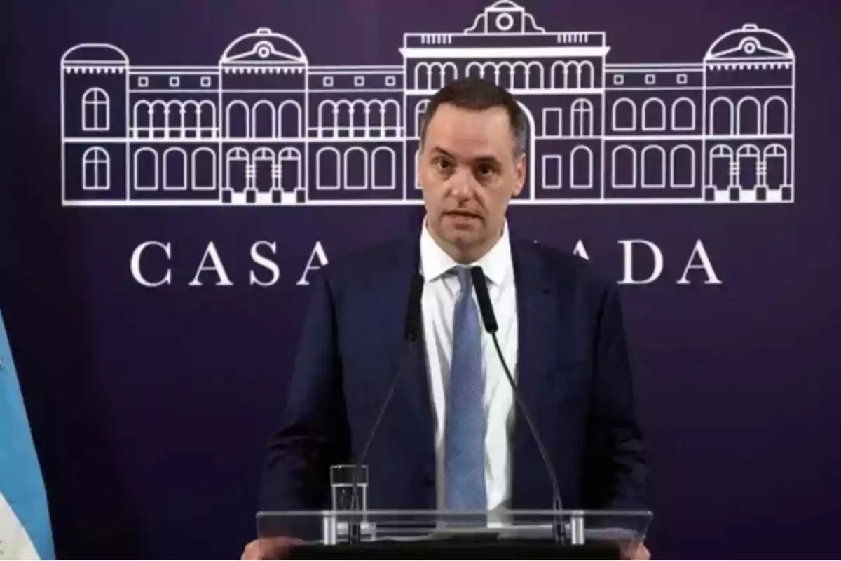 Hombre de traje hablando en un podio con micrófonos frente a un fondo que muestra un dibujo de un edificio y la palabra CASA