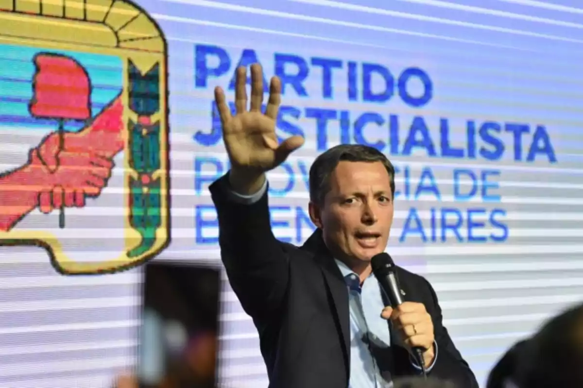 Un hombre hablando en un micrófono con el logo del Partido Justicialista de la Provincia de Buenos Aires de fondo.