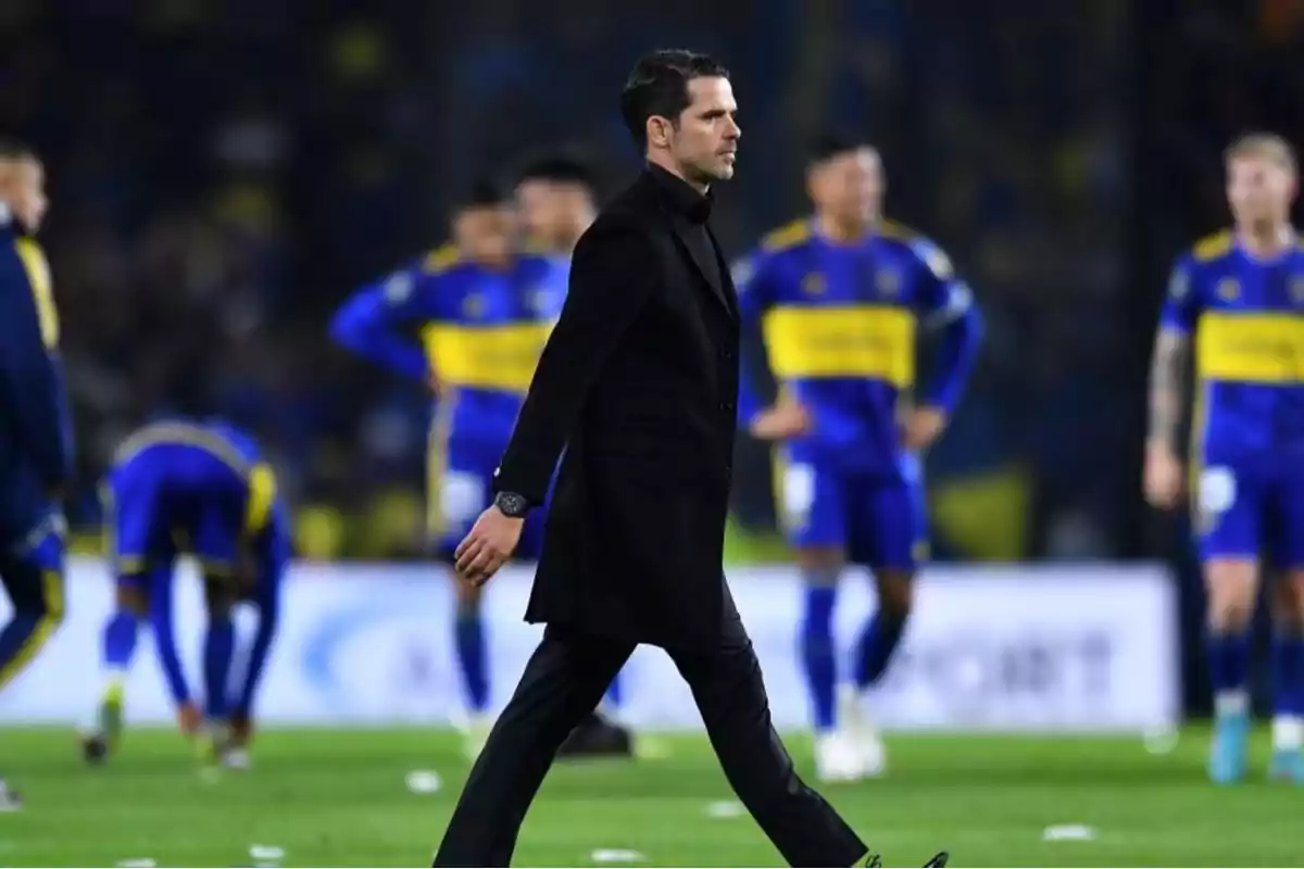 A man dressed in black walks across the soccer field while players in blue and yellow uniforms are in the background. A man dressed in black walks across the soccer field while players in blue and yellow uniforms are in the background.