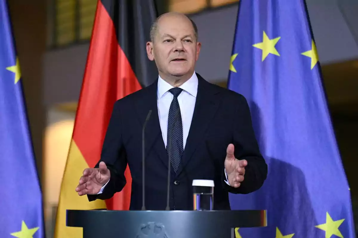Un hombre en traje hablando en un podio con las banderas de Alemania y la Unión Europea detrás.