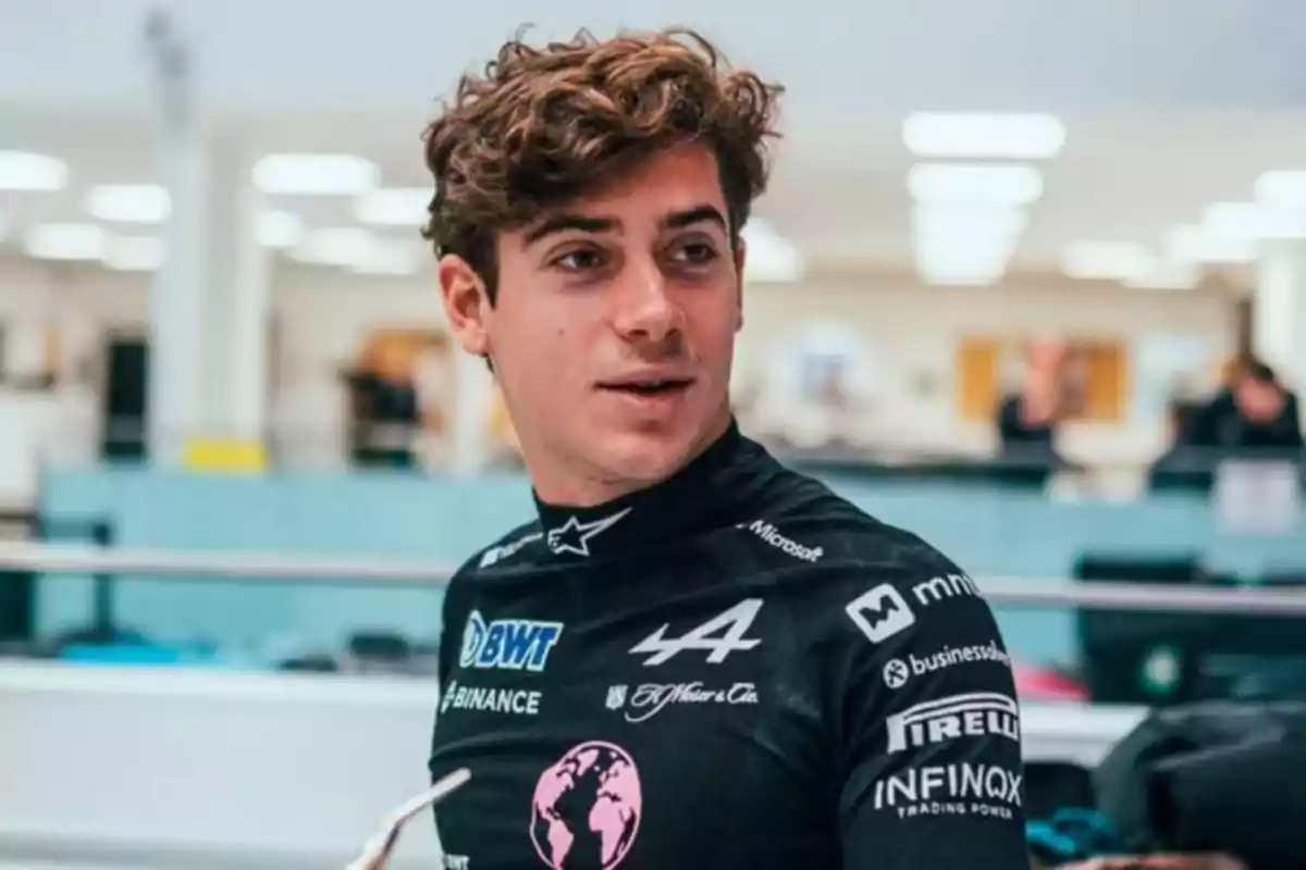 A man with curly hair is wearing a black racing suit with various logos in an indoor setting.