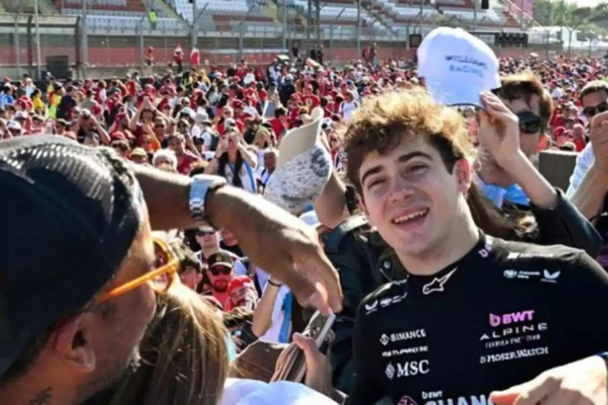 A smiling young man in a racing jersey is surrounded by a crowd at an outdoor event, while someone takes his picture. A smiling young man in a racing jersey is surrounded by a crowd at an outdoor event, while someone takes his picture.