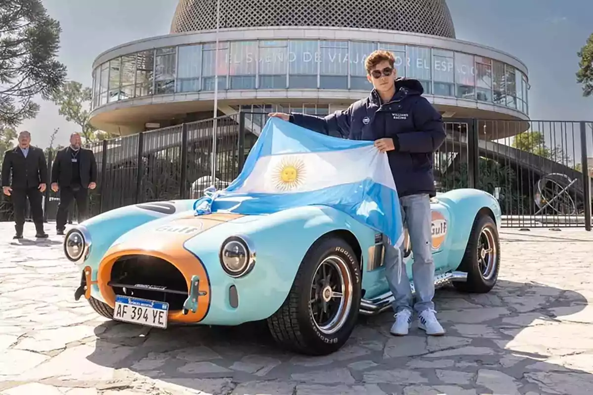 Young man with sunglasses holds an Argentine flag in front of a light blue and orange sports car parked, with a modern building in the background.