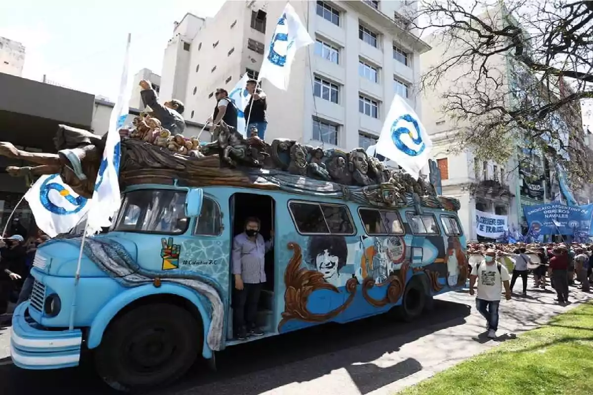 Un colectivo antiguo pintado de azul con murales y esculturas en el techo avanza por una calle durante una manifestación con personas portando banderas blancas y azules