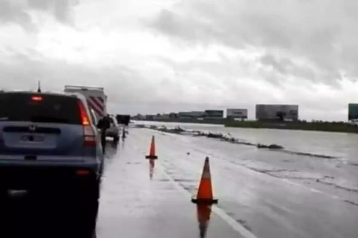 Wet road with orange cones and traffic under a cloudy sky.