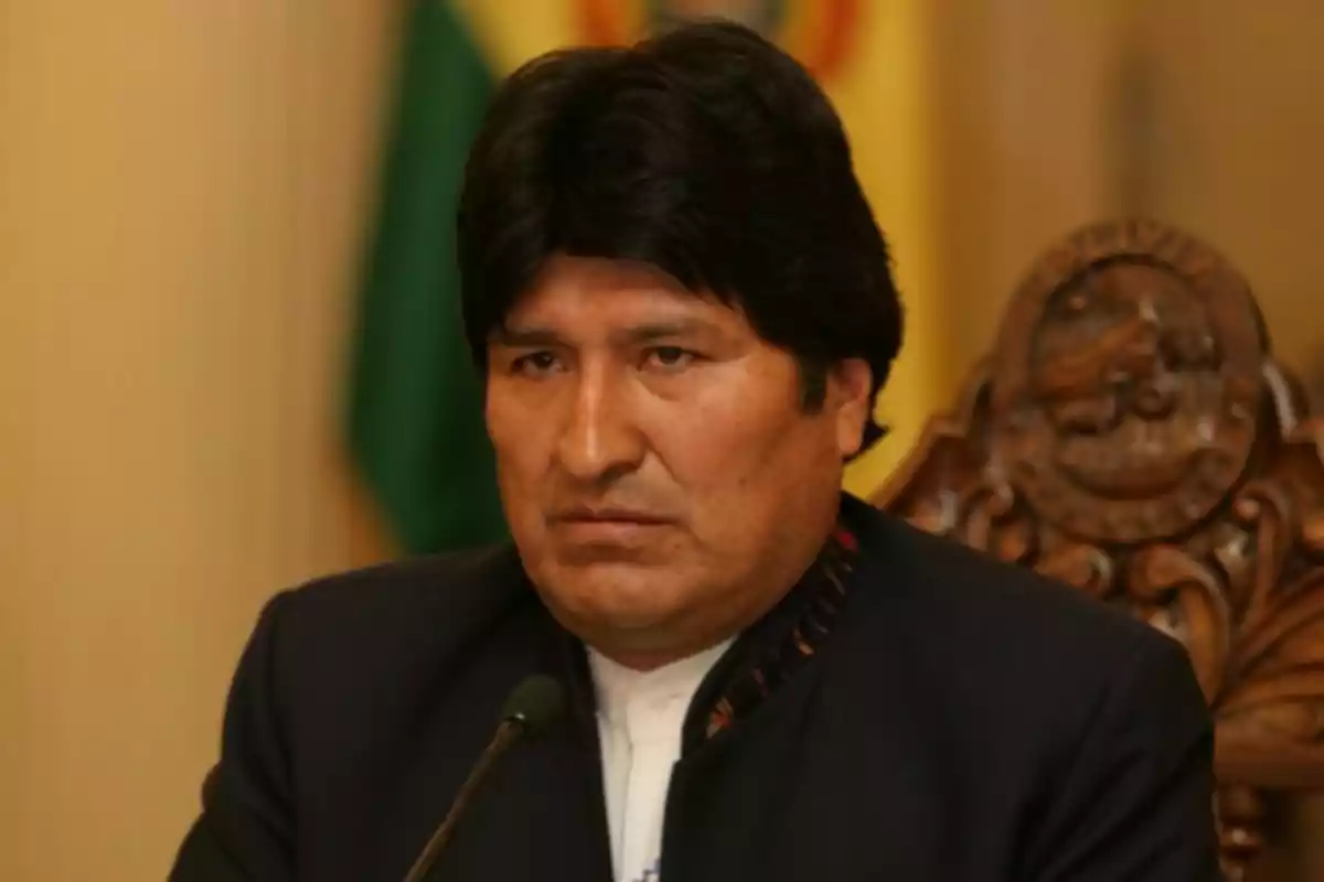 Un hombre con cabello oscuro y expresión seria sentado en una silla tallada con un fondo de bandera.