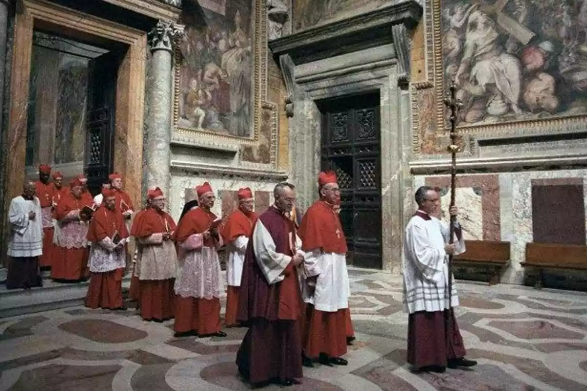 A group of cardinals dressed in red cassocks walks in procession inside a building with walls decorated with religious art. A group of cardinals dressed in red cassocks walks in procession inside a building with walls decorated with religious art.