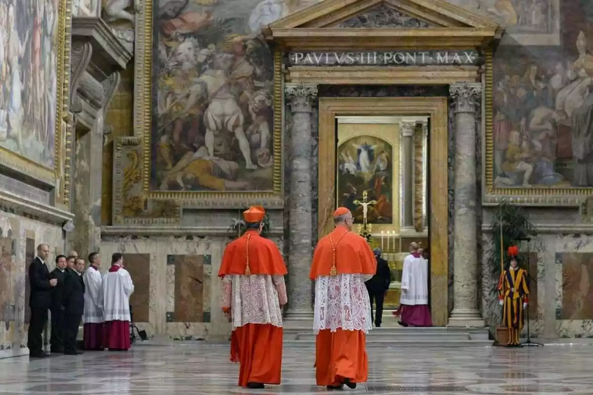 Two cardinals in red vestments walk down a hallway decorated with frescoes and columns, while several people watch from the side. Two cardinals in red vestments walk down a hallway decorated with frescoes and columns, while several people watch from the side.