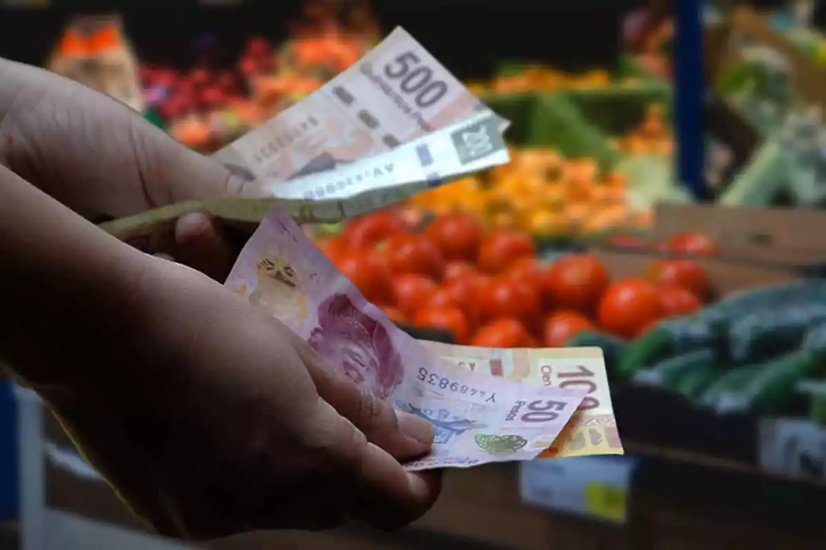 Una persona sostiene billetes de pesos mexicanos frente a un puesto de frutas y verduras en un mercado.