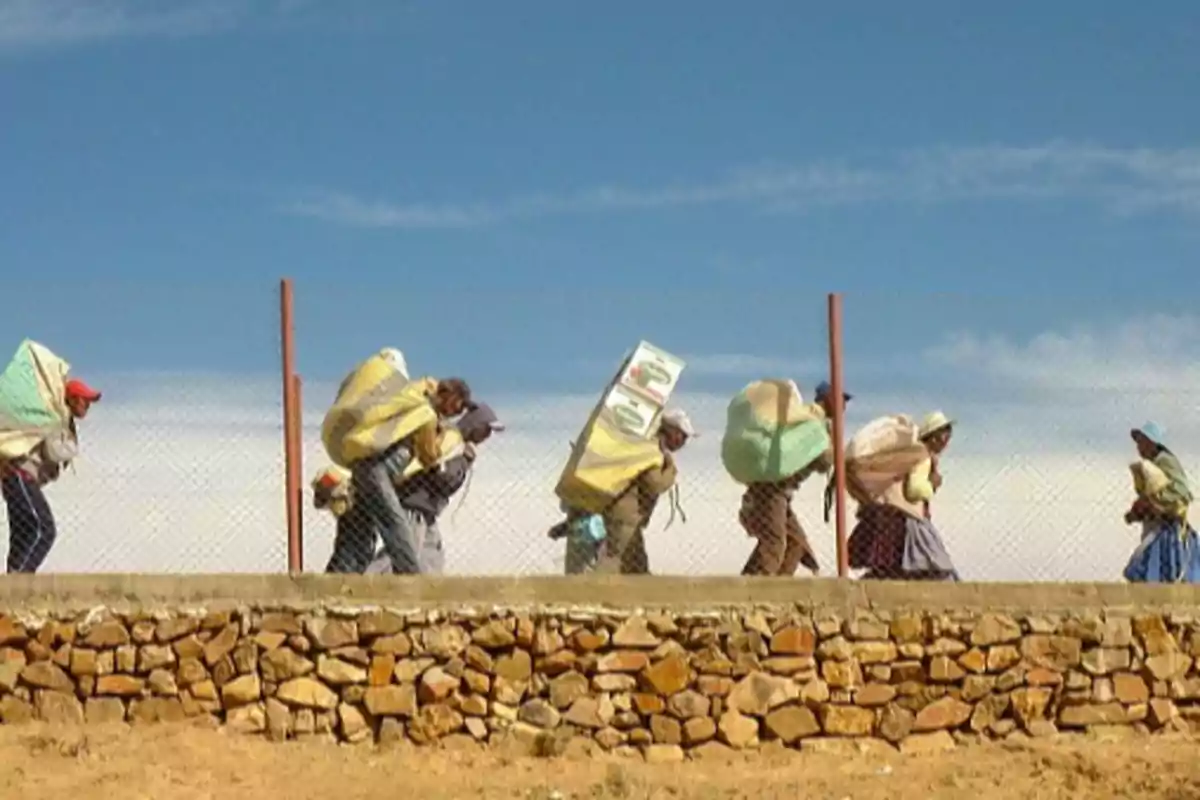 Un grupo de personas camina cargando grandes bultos sobre sus espaldas junto a una cerca de alambre bajo un cielo despejado.