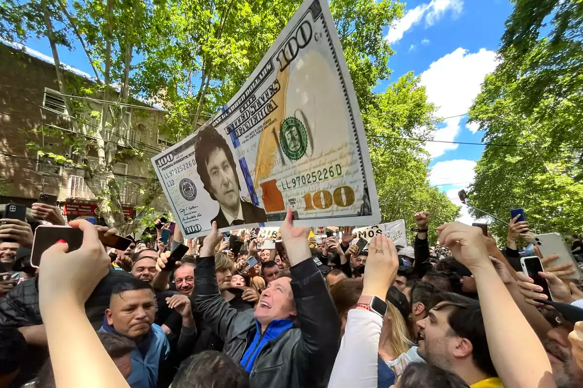 Una multitud de personas al aire libre sostiene un cartel en forma de billete de cien dólares con el rostro de un hombre en lugar del retrato habitual, mientras muchos toman fotos con sus teléfonos. Una multitud de personas al aire libre sostiene un cartel en forma de billete de cien dólares con el rostro de un hombre en lugar del retrato habitual, mientras muchos toman fotos con sus teléfonos.