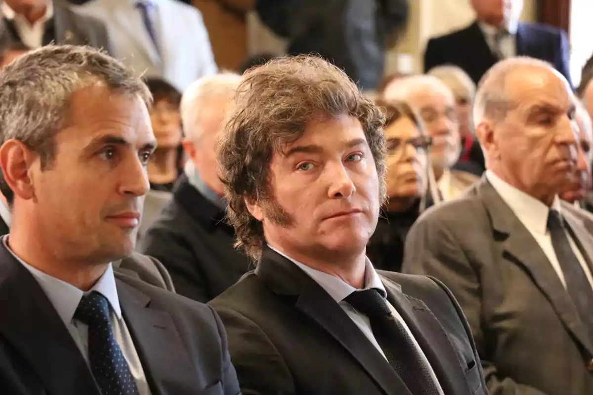 Men dressed in suits sitting at a formal event with several people in the background paying attention