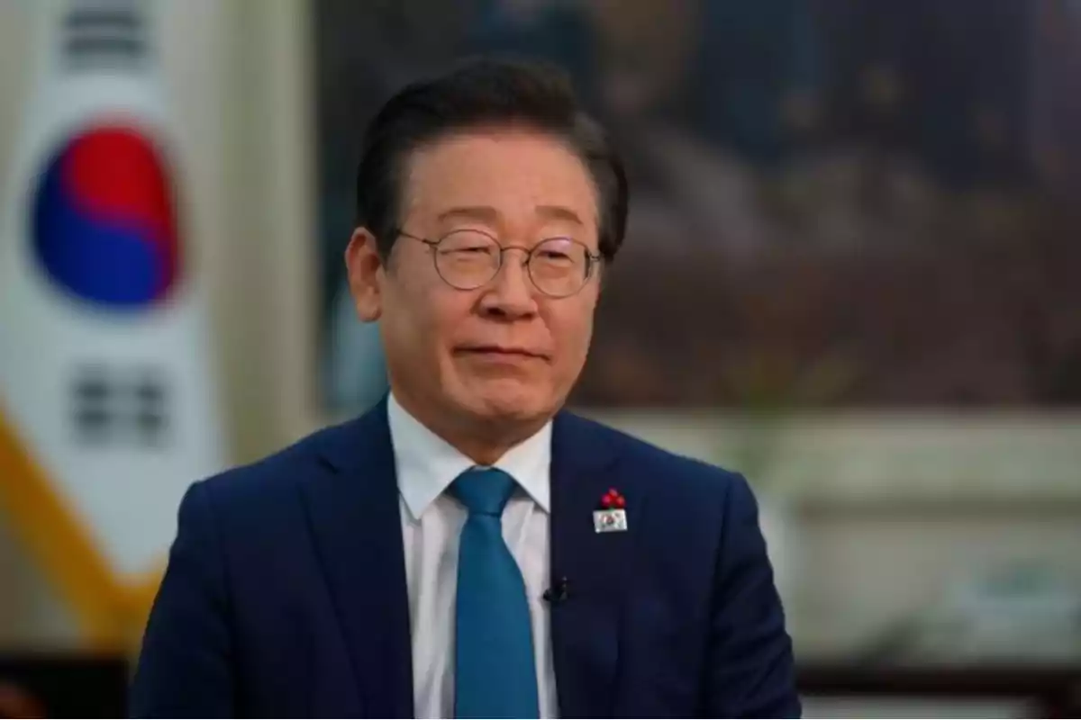 A man with glasses and a blue suit is sitting in front of a South Korean flag.