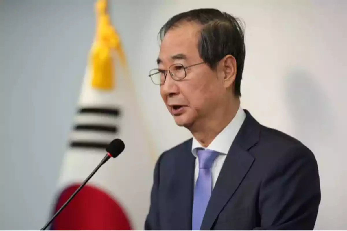 A man in a suit and tie speaks in front of a microphone with a South Korean flag in the background.