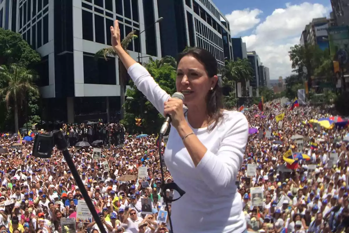 Una persona hablando en un mitin frente a una multitud en una ciudad con edificios altos y cielo despejado.