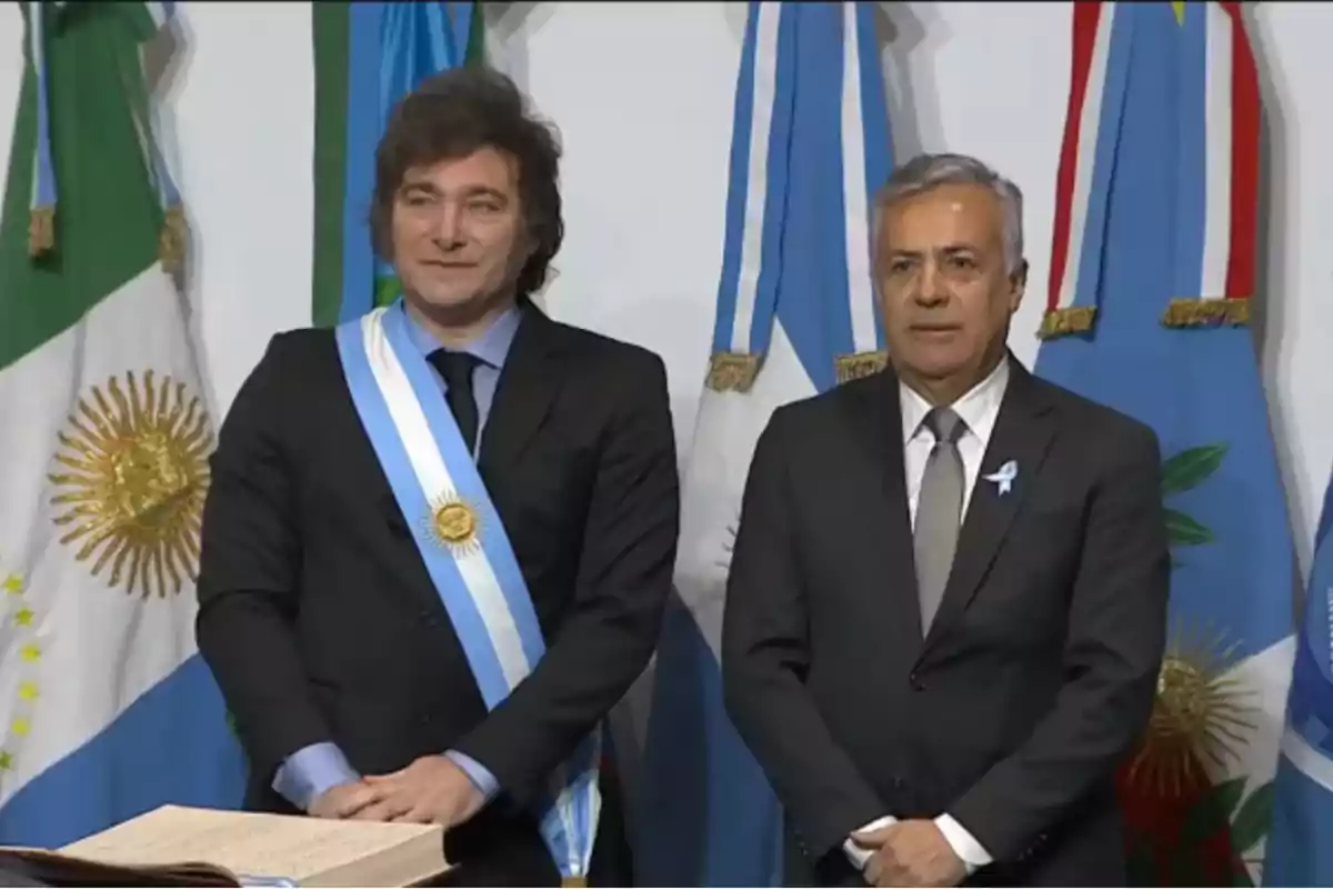 Dos hombres de traje posan frente a varias banderas, uno de ellos lleva una banda presidencial y una medalla.