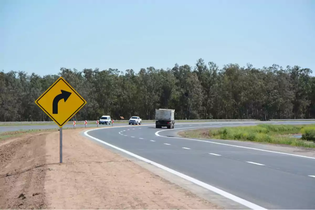 Señal de tráfico indicando una curva a la derecha en una carretera con vehículos circulando y árboles al fondo.