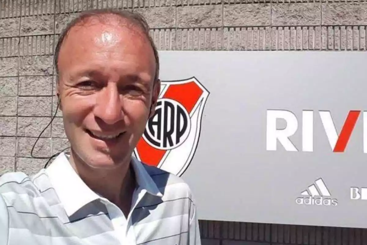 A smiling man takes a selfie in front of a sign with a soccer club's emblem and the word A smiling man takes a selfie in front of a sign with a soccer club's emblem and the word