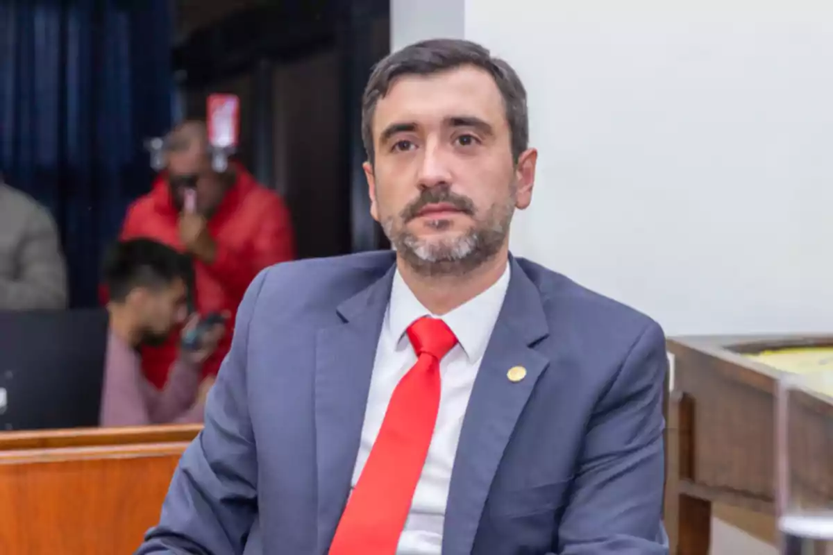 Hombre de cabello corto y barba con traje azul y corbata roja sentado en una oficina