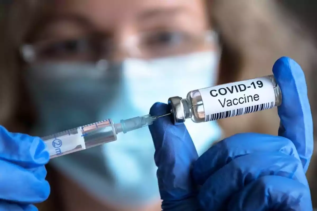A person with blue gloves holds a syringe and a vial labeled as COVID-19 vaccine.