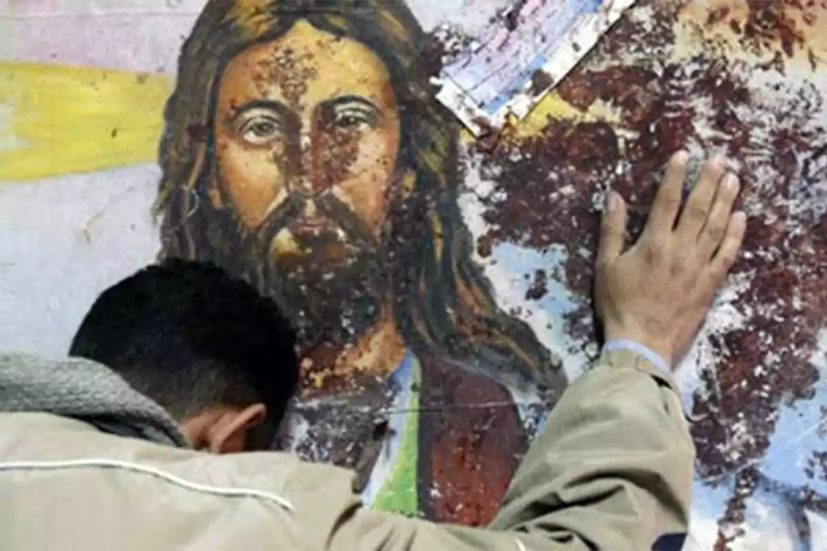 Un hombre con la cabeza inclinada y la mano apoyada sobre una imagen religiosa dañada.