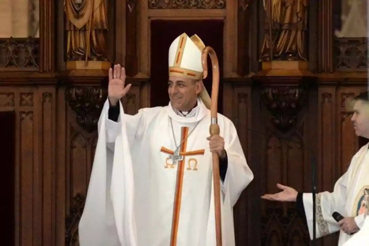 Un obispo vestido con vestimentas litúrgicas blancas y una mitra, sosteniendo un báculo, saluda con la mano levantada en un entorno eclesiástico decorado con tallas de madera.