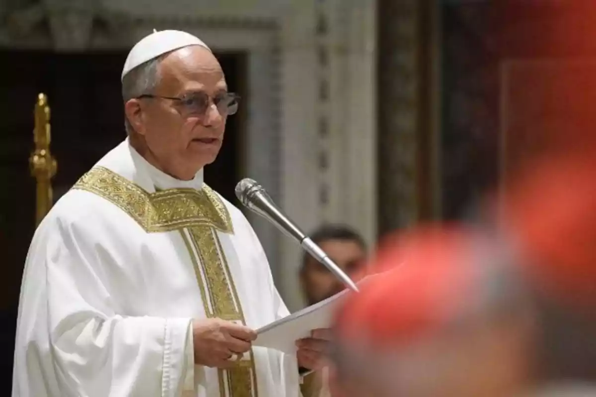 Un clérigo en vestimenta religiosa blanca y dorada habla frente a un micrófono en un entorno eclesiástico. Un clérigo en vestimenta religiosa blanca y dorada habla frente a un micrófono en un entorno eclesiástico.