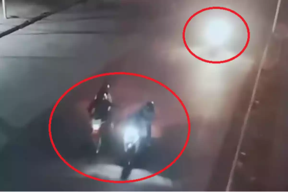 Dos motocicletas circulando por una carretera de noche con luces brillantes en el fondo.