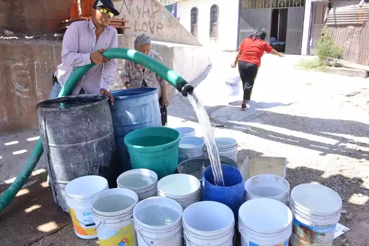 People filling buckets and barrels with water from a large hose in an urban area under the sun